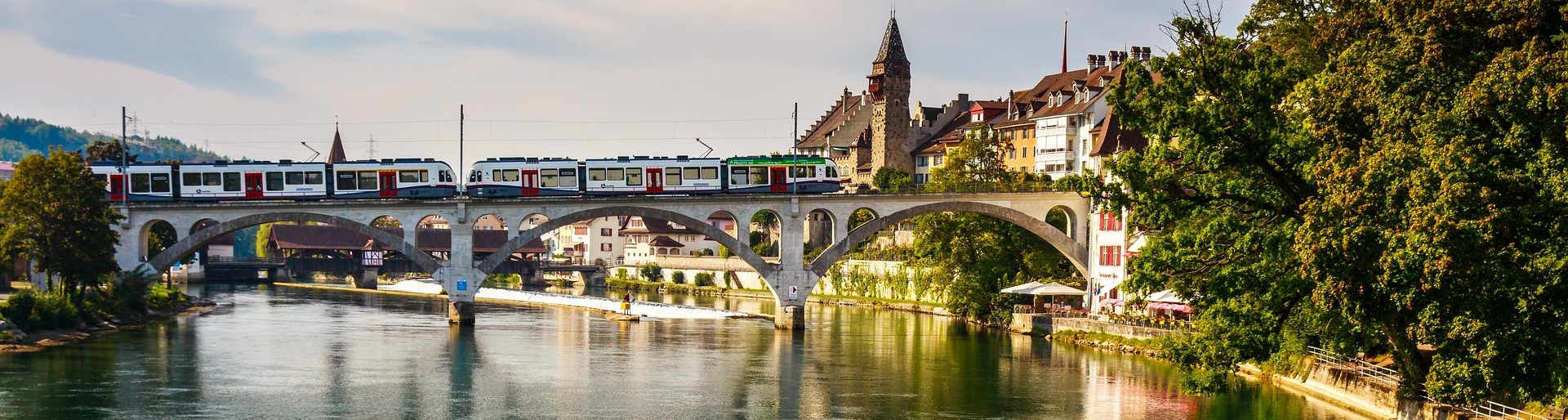 Ausflugsziele im Aargau - Blick auf eine Stadt im Aargau mit einem Fluss, der hindurchfließt, und einer Straßenbahn, die auf der Brücke über dem Fluss fährt