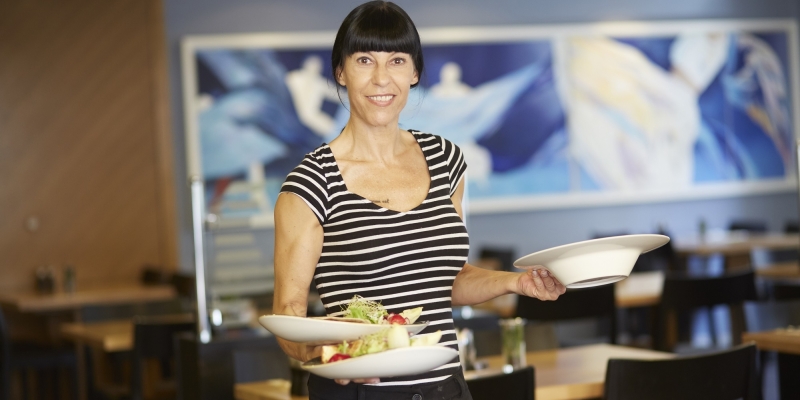 Eine schwarzhaarige, lächelnde Servicekraft hält 3 Teller in ihren Händen.