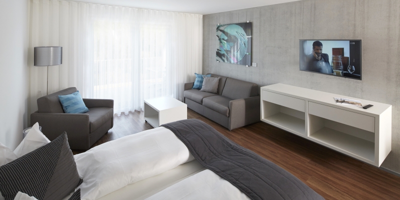Modernes Hotelzimmer mit Doppelbett, grauem Sofa, einem Fernseher und bodentiefen Fenstern